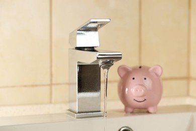 Photo of Water saving concept. Stream of water flowing from tap and piggy bank indoors, selective focus