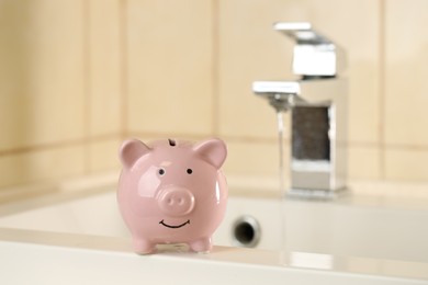 Photo of Water saving concept. Piggy bank on sink and stream of water flowing from tap indoors, selective focus