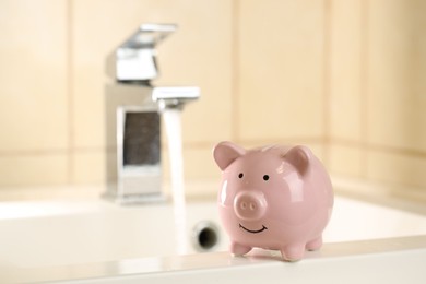 Photo of Water saving concept. Piggy bank on sink and stream of water flowing from tap indoors, selective focus