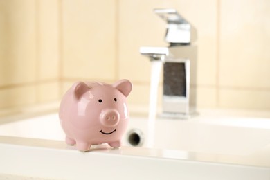 Photo of Water saving concept. Piggy bank on sink and stream of water flowing from tap indoors, selective focus