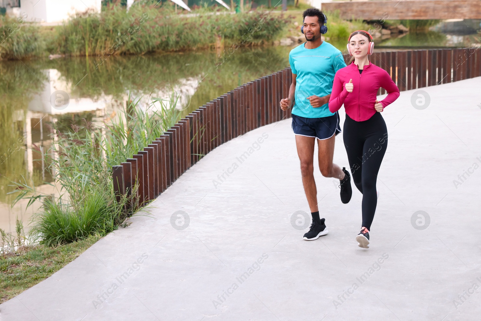 Photo of Sporty couple in headphones running near river outdoors. Space for text