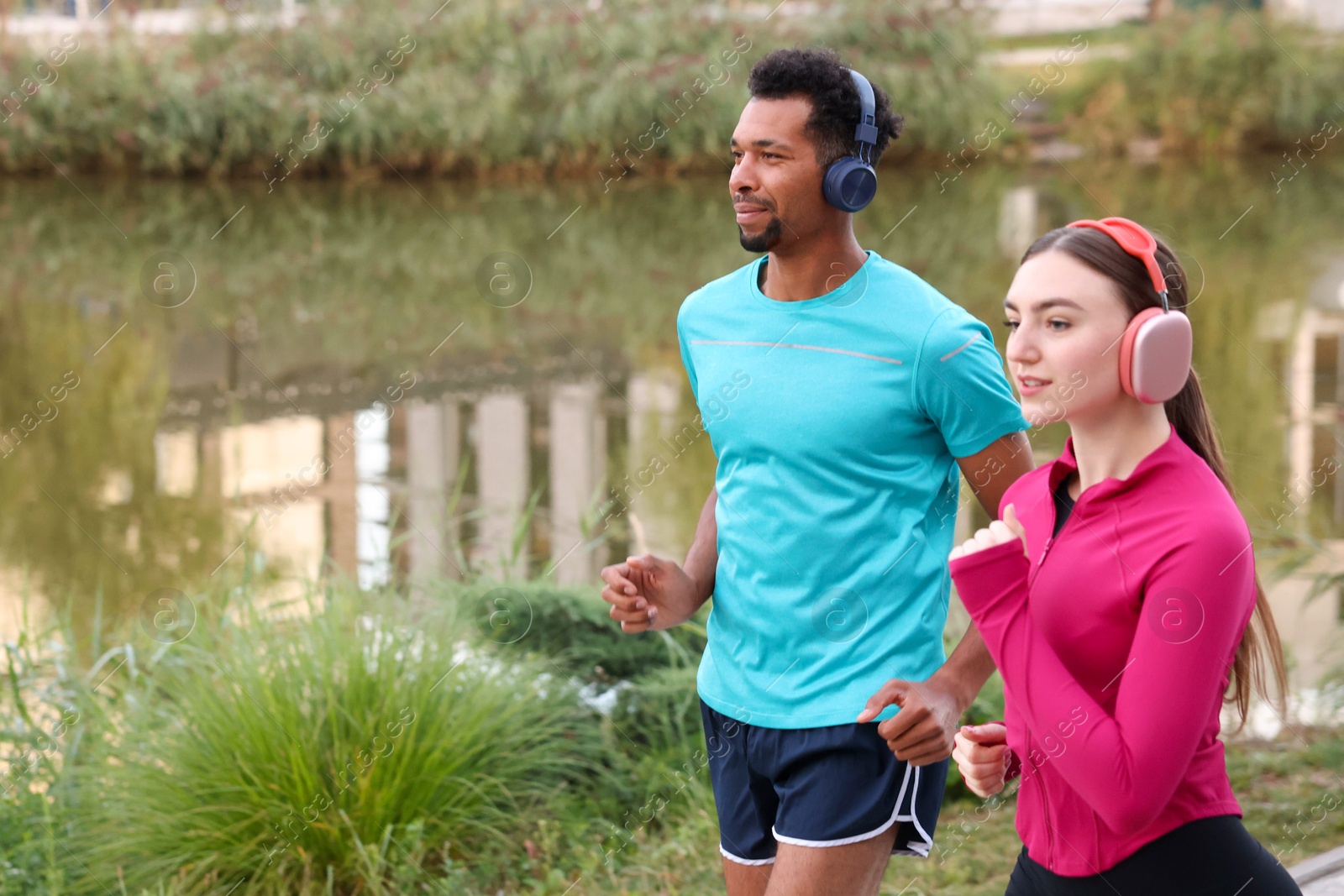 Photo of Sporty couple in headphones running near river outdoors. Space for text