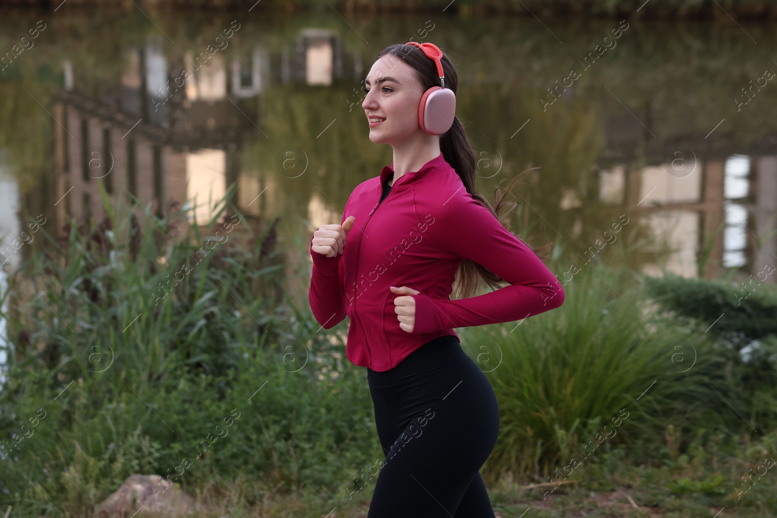 Smiling woman in headphones running near river outdoors Photo of Smiling woman in headphones running near river outdoors