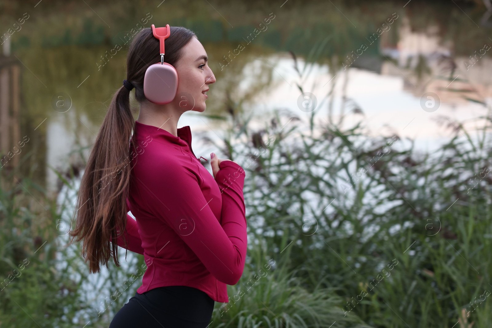Photo of Smiling woman in headphones running near river outdoors. Space for text