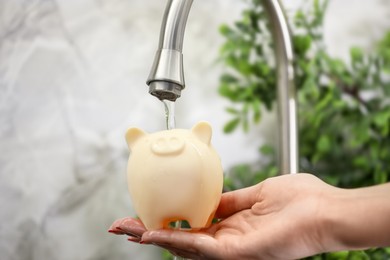 Photo of Water saving concept. Woman holding piggy bank under tap indoors, closeup