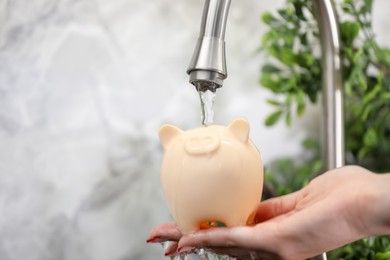 Photo of Water saving concept. Woman holding piggy bank under tap indoors, closeup