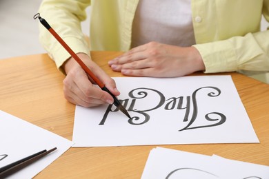 Photo of Calligraphy. Woman with brush writing words The End on sheet of paper at wooden table indoors, closeup