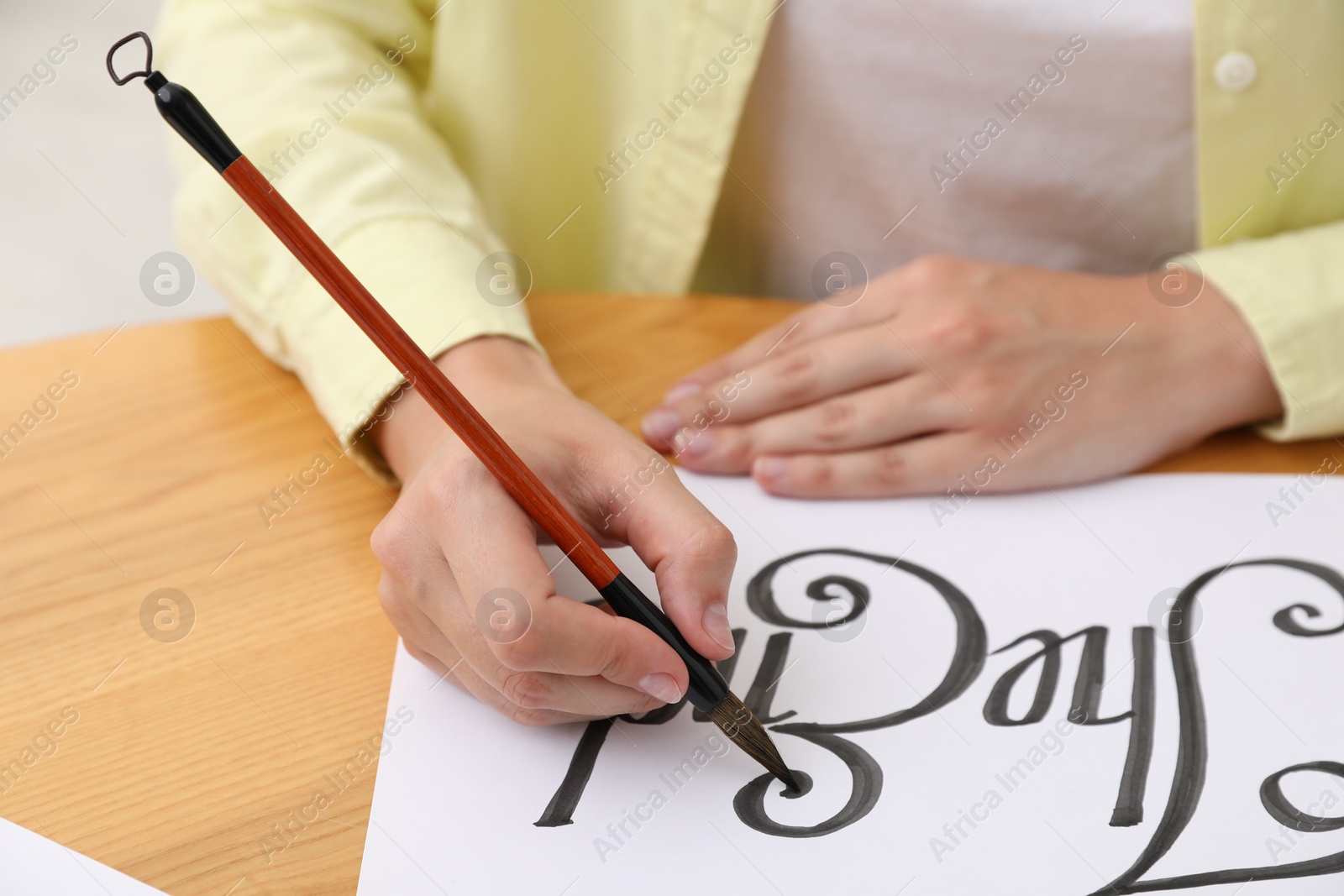 Photo of Calligraphy. Woman with brush writing words The End on sheet of paper at wooden table indoors, closeup