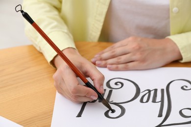 Photo of Calligraphy. Woman with brush writing words The End on sheet of paper at wooden table indoors, closeup