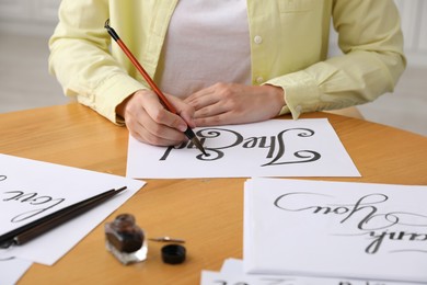 Photo of Calligraphy. Woman with brush writing words The End on sheet of paper at wooden table indoors, closeup