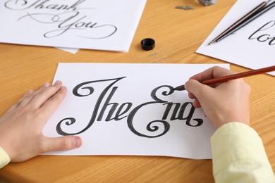 Photo of Calligraphy. Woman with brush writing words The End on sheet of paper at wooden table, closeup