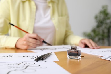 Photo of Calligraphy. Woman with brush writing at wooden table indoors, focus on inkwell and fountain pens