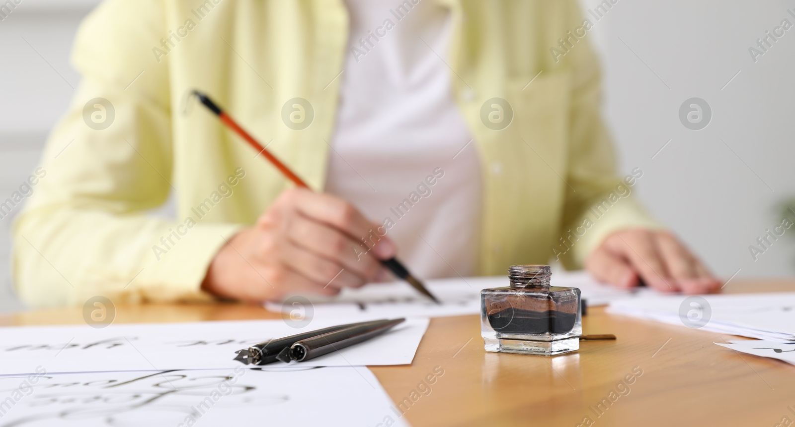 Calligraphy. Woman with brush writing at wooden table indoors, focus on inkwell and fountain pens Photo of Calligraphy. Woman with brush writing at wooden table indoors, focus on inkwell and fountain pens