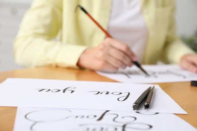 Photo of Calligraphy. Woman with brush writing at table indoors, focus on fountain pens and sheets of paper