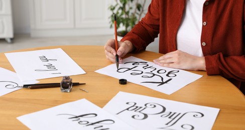 Photo of Calligraphy. Woman with brush writing phrase Keep Going on sheet of paper at wooden table indoors, closeup