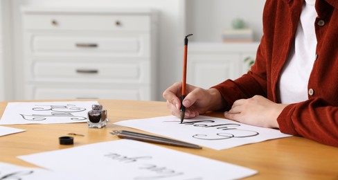 Photo of Calligraphy. Woman with brush writing word Enjoy on sheet of paper at wooden table, closeup