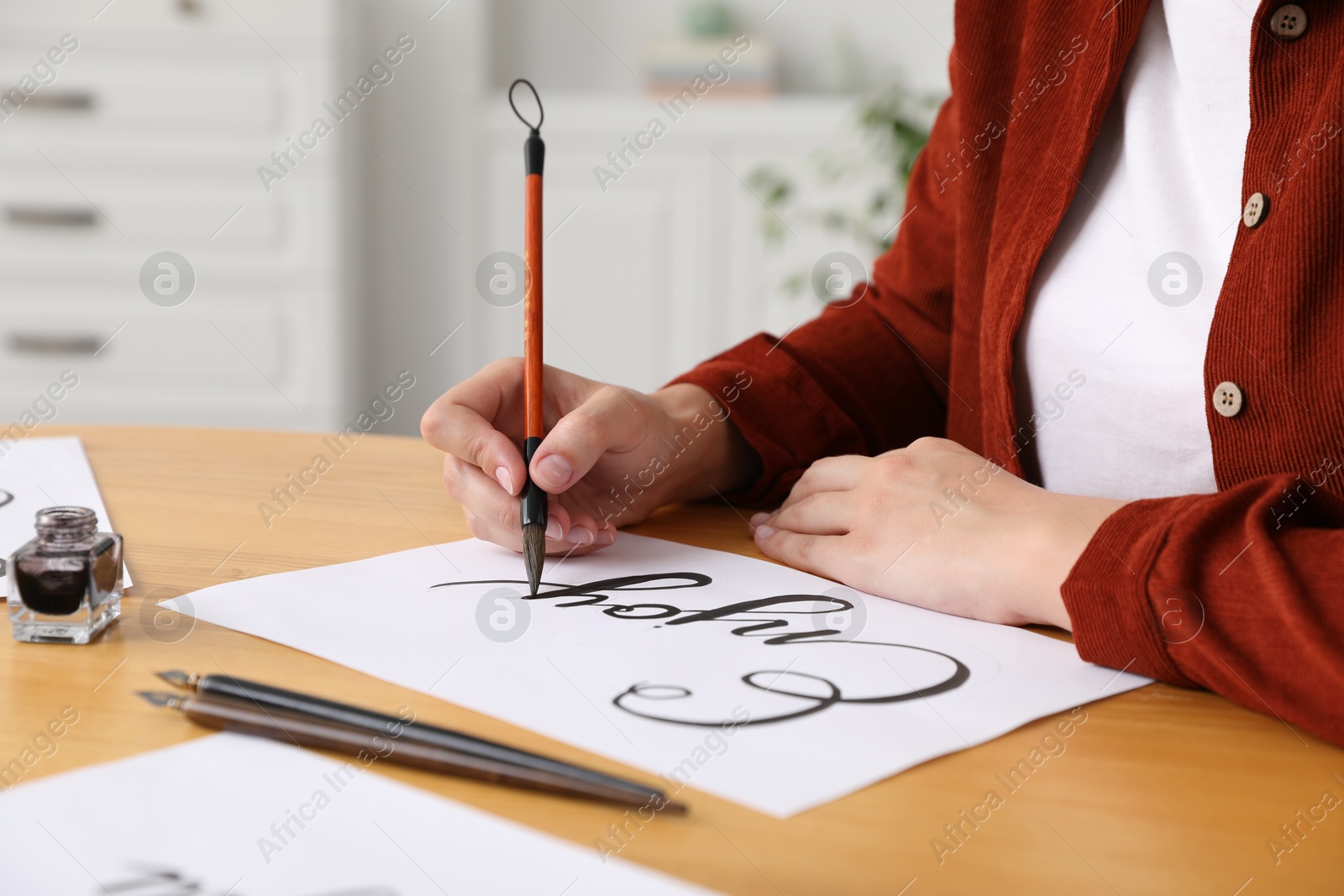 Calligraphy. Woman with brush writing word Enjoy on sheet of paper at wooden table, closeup Photo of Calligraphy. Woman with brush writing word Enjoy on sheet of paper at wooden table, closeup