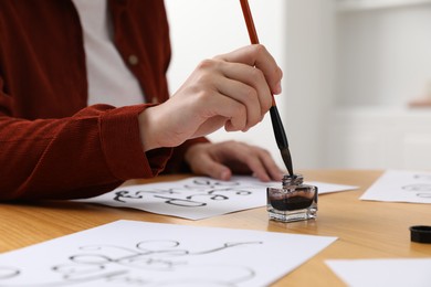 Photo of Calligraphy. Woman dipping brush into inkwell at wooden table indoors, closeup