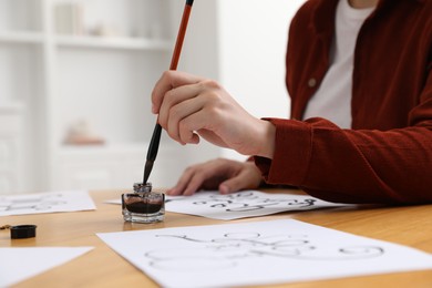 Photo of Calligraphy. Woman dipping brush into inkwell at wooden table indoors, closeup