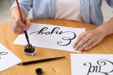 Photo of Calligraphy. Woman dipping brush into inkwell at wooden table indoors, closeup