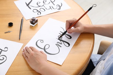 Photo of Calligraphy. Woman with brush writing word Enjoy on sheet of paper at wooden table indoors, closeup