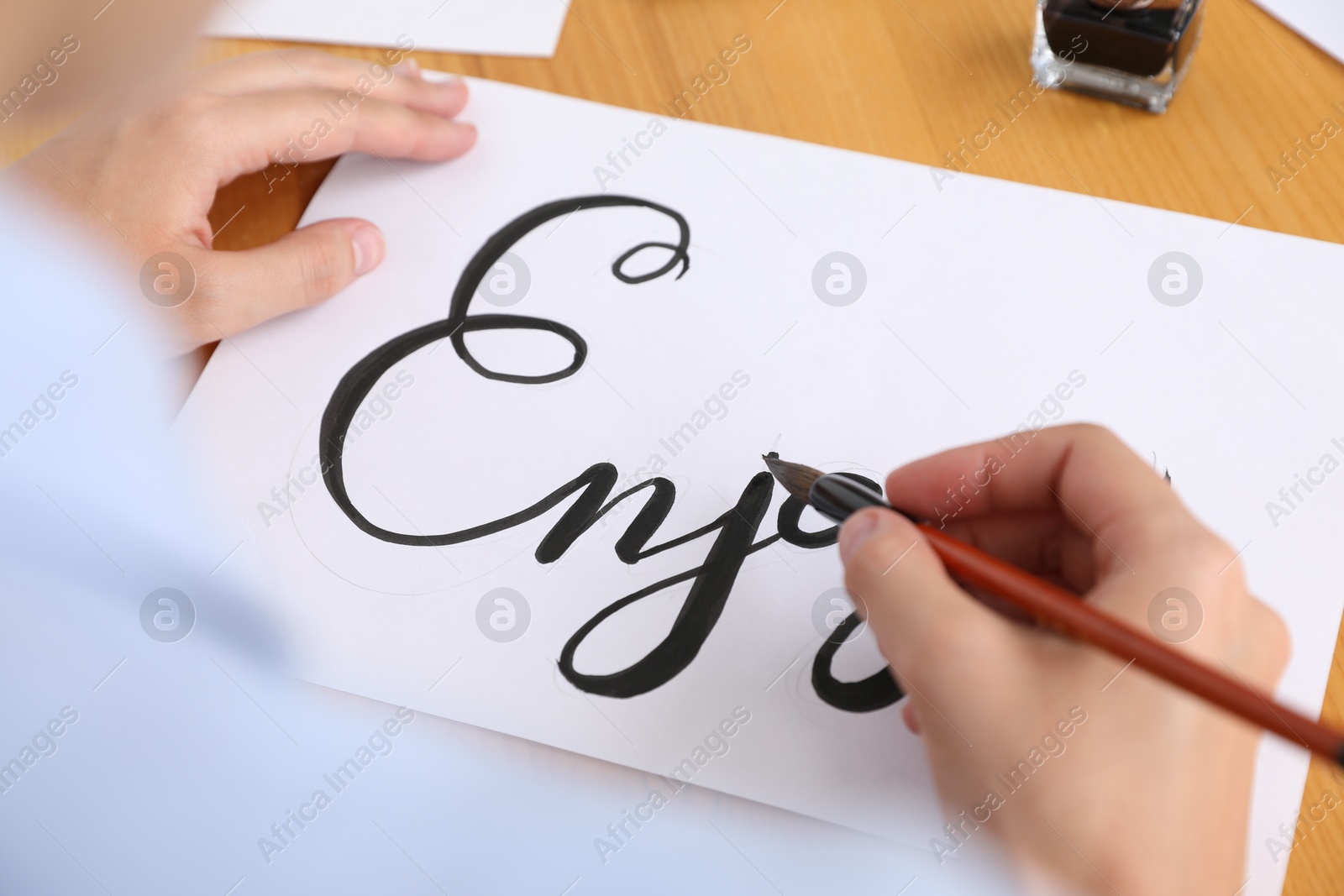 Calligraphy. Woman with brush writing word Enjoy on sheet of paper at wooden table, above view Photo of Calligraphy. Woman with brush writing word Enjoy on sheet of paper at wooden table, above view
