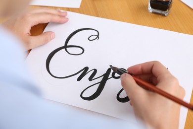 Photo of Calligraphy. Woman with brush writing word Enjoy on sheet of paper at wooden table, above view