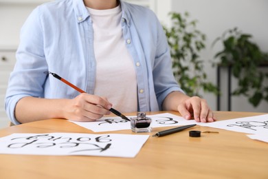 Photo of Calligraphy. Woman with brush writing word Enjoy on sheet of paper at wooden table indoors, closeup