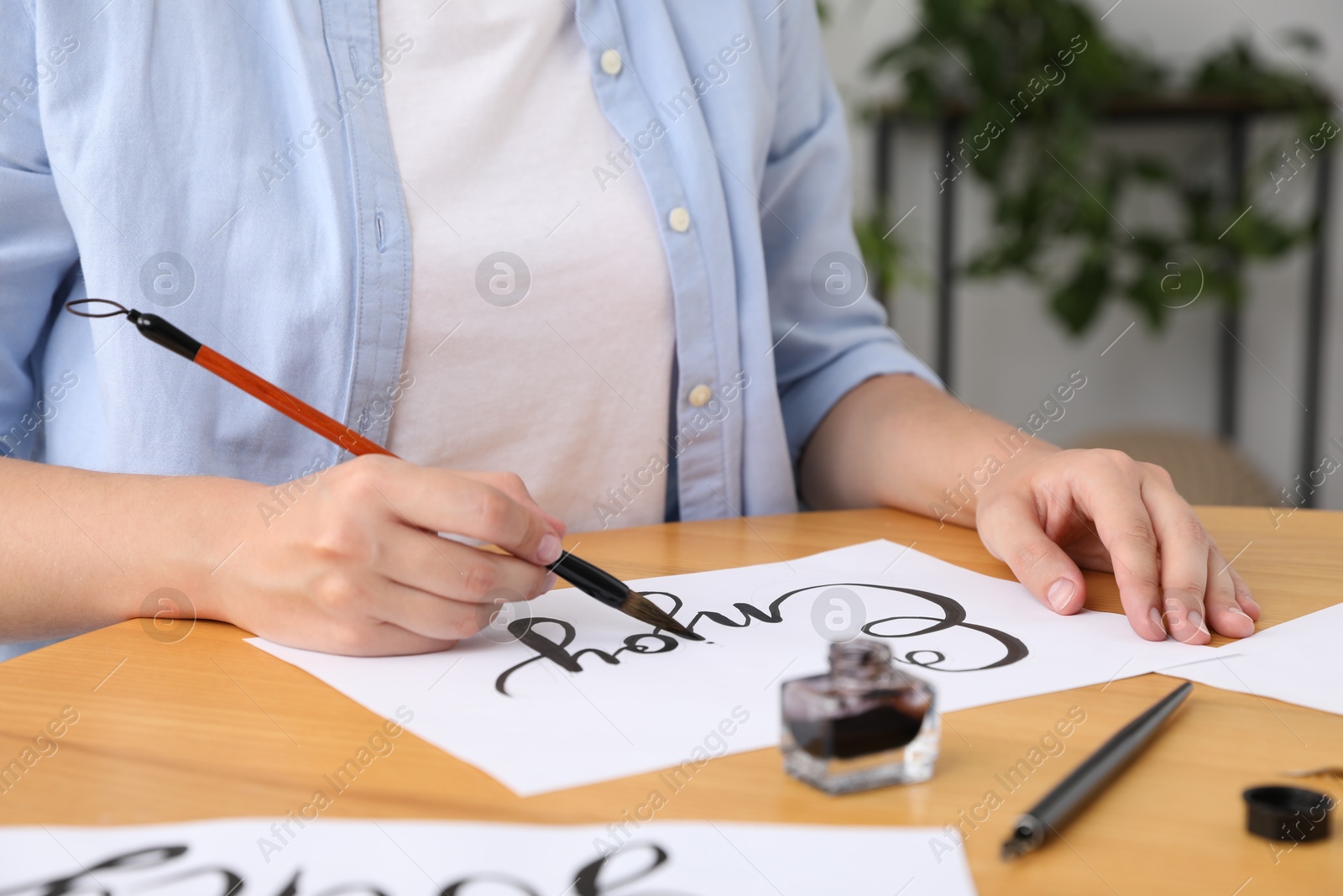 Calligraphy. Woman with brush writing word Enjoy on sheet of paper at wooden table indoors, closeup Photo of Calligraphy. Woman with brush writing word Enjoy on sheet of paper at wooden table indoors, closeup