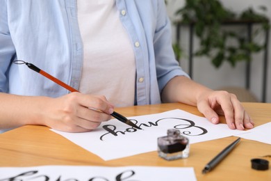 Photo of Calligraphy. Woman with brush writing word Enjoy on sheet of paper at wooden table indoors, closeup