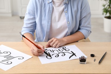 Photo of Calligraphy. Woman with brush writing phrase Keep Going on sheet of paper at wooden table indoors, closeup