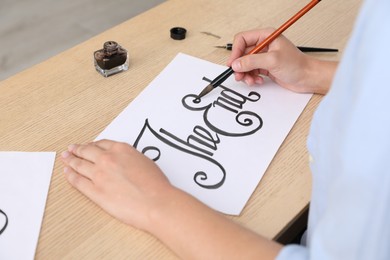 Photo of Calligraphy. Woman with brush writing words The End on sheet of paper at wooden table, closeup