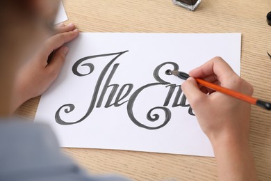 Photo of Calligraphy. Woman with brush writing words The End on sheet of paper at wooden table, above view