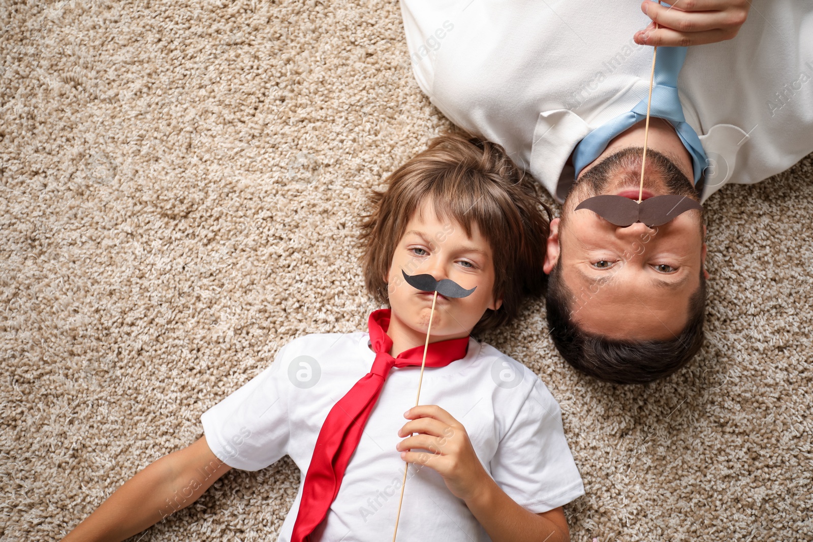 Father and son with paper moustache lying on rug, top view. Space for text Photo of Father and son with paper moustache lying on rug, top view. Space for text