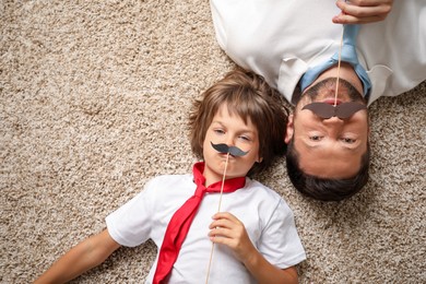 Photo of Father and son with paper moustache lying on rug, top view. Space for text