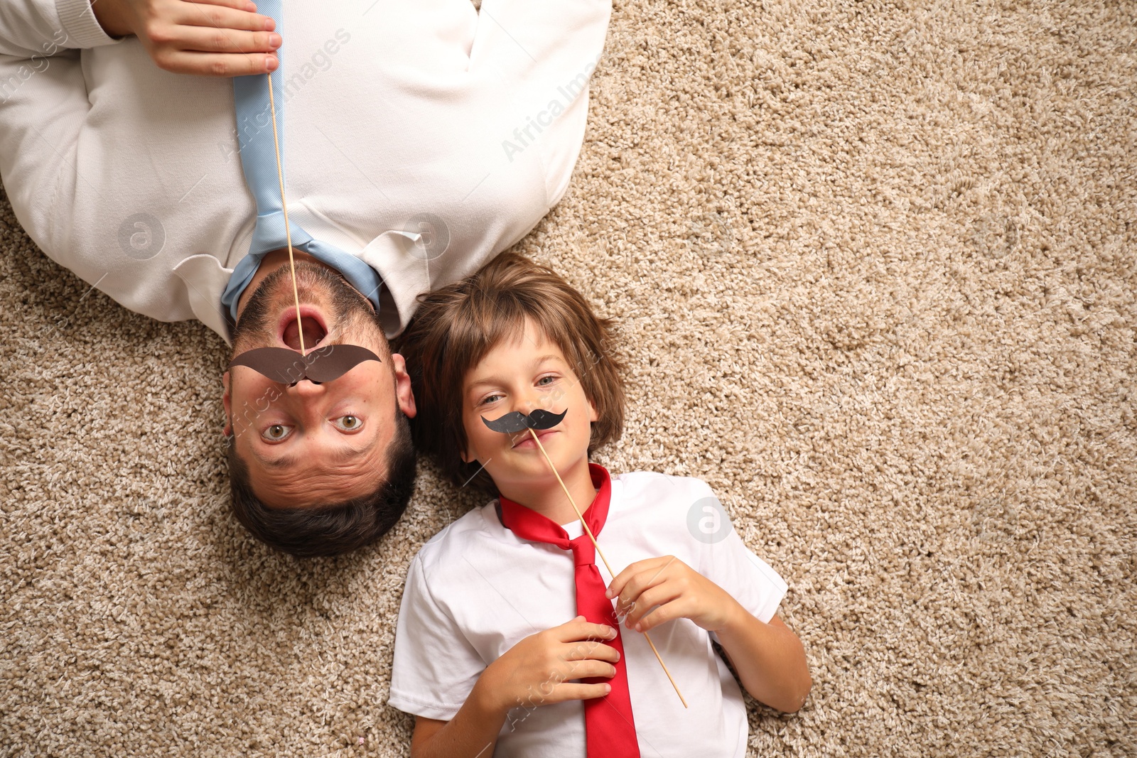 Photo of Emotional father and son with paper moustache lying on rug, top view. Space for text