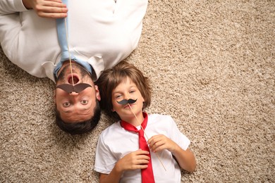 Photo of Emotional father and son with paper moustache lying on rug, top view. Space for text