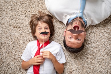 Photo of Emotional father and son with paper moustache lying on rug, top view