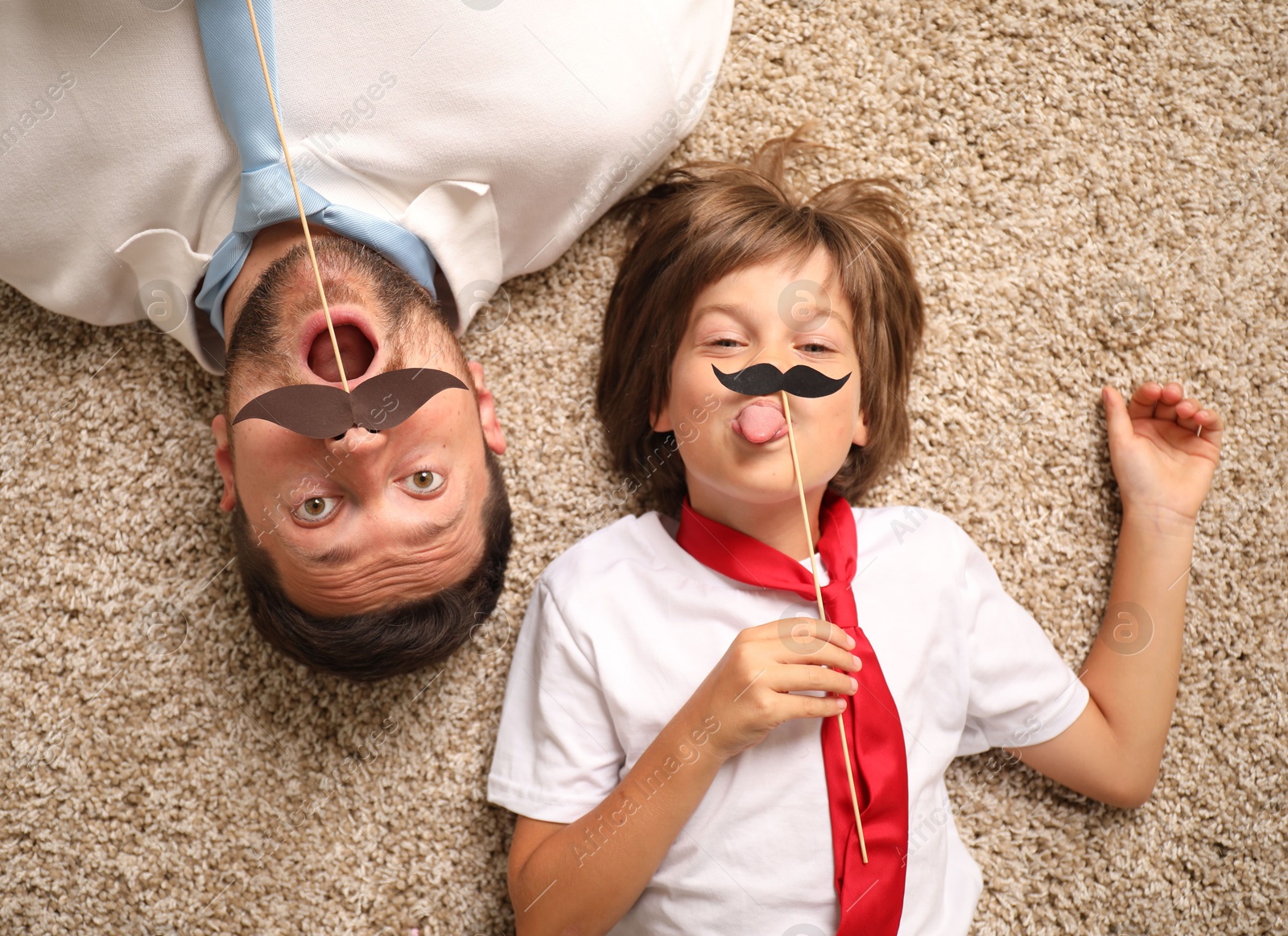 Photo of Emotional father and son with paper moustache lying on rug, top view