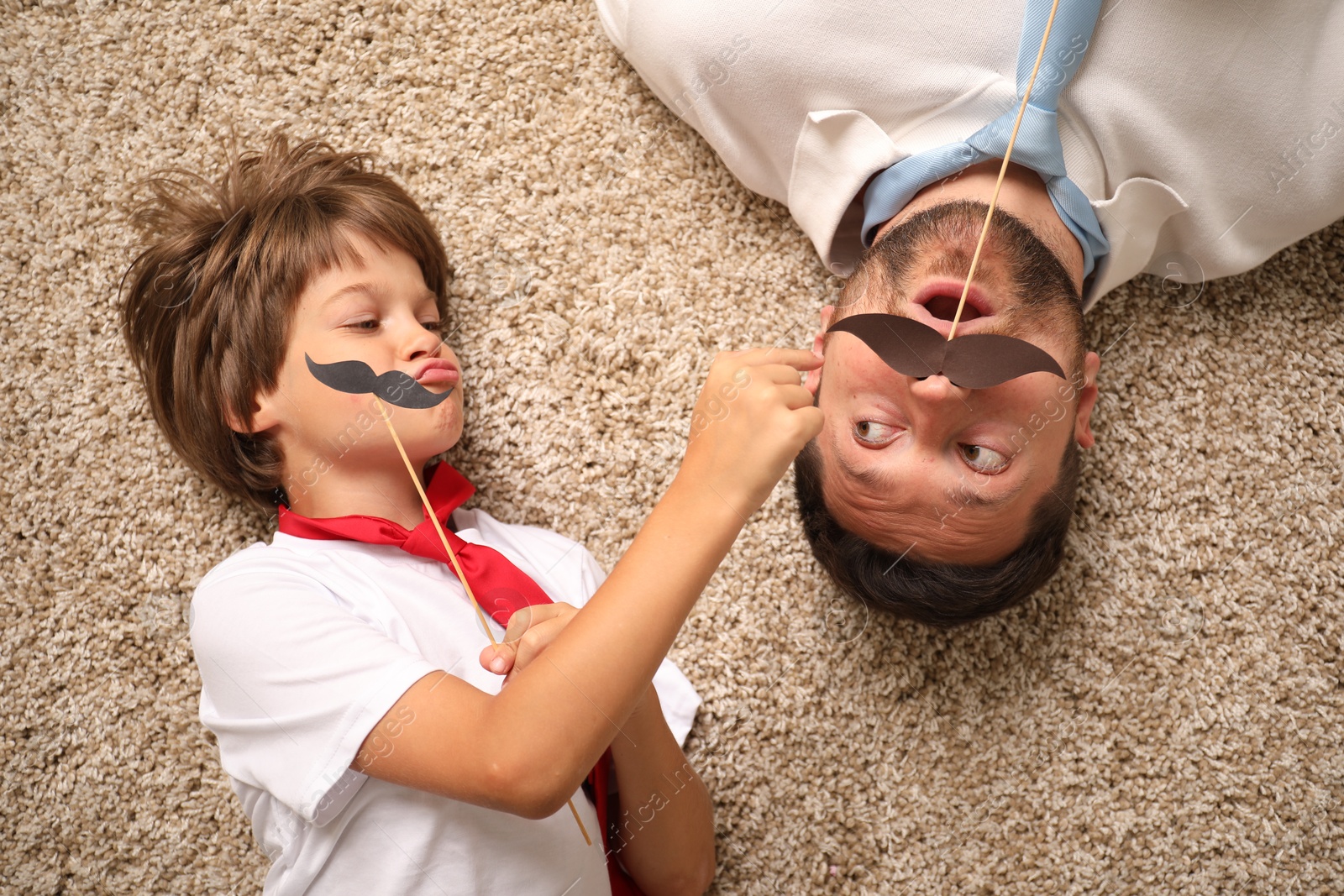 Photo of Emotional father and son with paper moustache lying on rug, top view