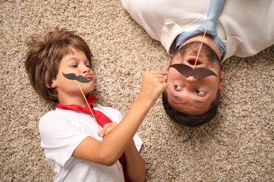 Photo of Emotional father and son with paper moustache lying on rug, top view