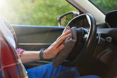 Photo of Man wiping steering wheel with grey microfiber rag in car salon, closeup