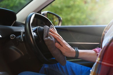 Photo of Man wiping steering wheel with grey microfiber rag in car salon, closeup
