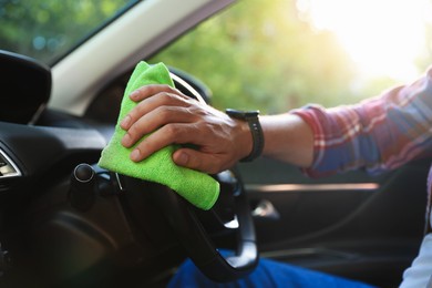 Photo of Man wiping steering wheel with green microfiber rag in car salon, closeup