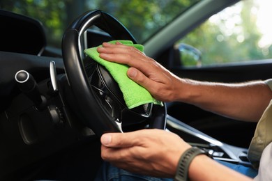 Photo of Man wiping steering wheel with green microfiber rag in car salon, closeup