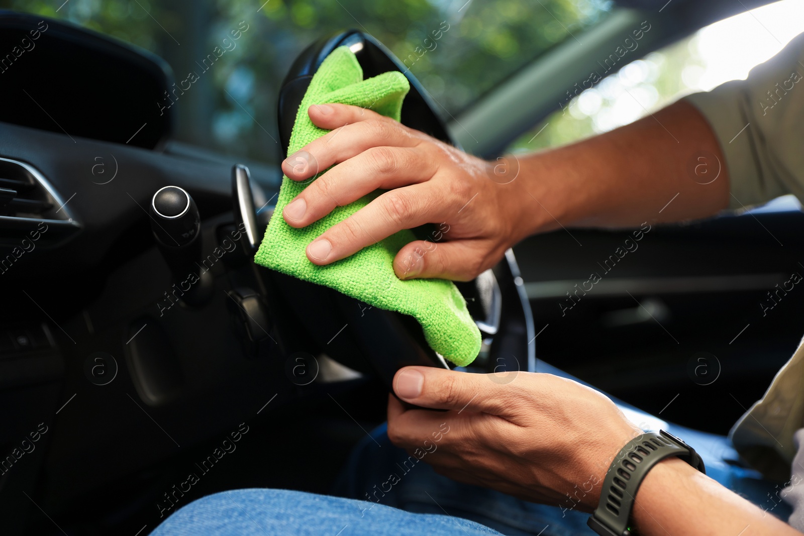 Photo of Man wiping steering wheel with green microfiber rag in car salon, closeup