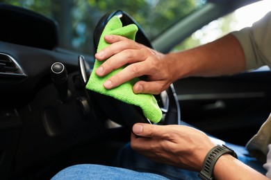 Photo of Man wiping steering wheel with green microfiber rag in car salon, closeup