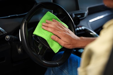 Photo of Man wiping steering wheel with green microfiber rag in car salon, closeup