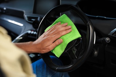 Photo of Man wiping steering wheel with green microfiber rag in car salon, closeup