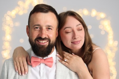 Photo of Making marriage proposal. Woman with engagement ring and her happy fiance hugging on light grey background with blurred lights, bokeh effect